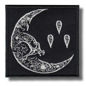 Può includere: Una toppa quadrata ricamata nera con un design a mezzaluna bianca caratterizzato da intricati motivi floreali all'interno della forma lunare, accompagnata da tre piccole forme bianche a goccia posizionate a destra della luna.