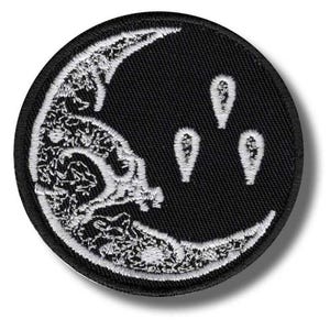 Könnte beinhalten: Ein kreisförmiger bestickter Aufnäher mit schwarzem Hintergrund und einem weißen Mondsichel-Design. Der Mond ist mit komplizierten weißen Mustern verziert und wird von drei kleinen, weißen, tropfenförmigen Symbolen auf der rechten Seite begleitet.