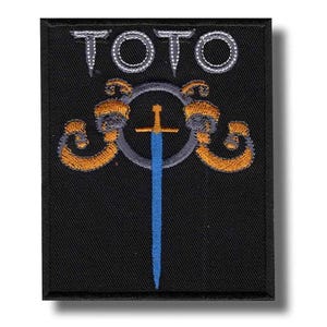 Könnte beinhalten: Ein rechteckiger, schwarz bestickter Aufnäher mit dem Wort TOTO in weißen Großbuchstaben am oberen Rand. Unter dem Text befindet sich ein grauer Kreisring, flankiert von zwei gold-orangen Schnörkelmustern und einem vertikalen blauen Schwert.