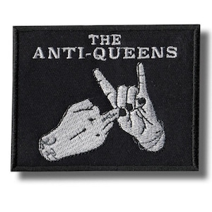 Op de afbeelding: Een rechthoekige zwarte geborduurde patch met de witte tekst THE ANTI-QUEENS bovenaan. Onder de tekst zijn twee handen in wit borduurwerk afgebeeld, gepositioneerd in een handgebaar met de vingers omhoog en naar buiten gericht.