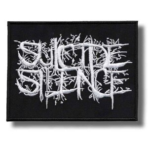 May include: A rectangular black fabric patch featuring white embroidered text that reads Suicide Silence. The lettering is stylized with intricate, branch-like extensions and sharp, jagged edges, creating a dense and complex visual design.
