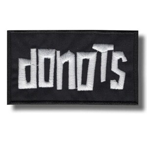 Puede incluir: Un parche rectangular negro bordado con el texto blanco "donots" en una fuente sans-serif negrita y cuadrada. El bordado tiene un patrón de puntadas texturizado visible en todas las letras, sobre un fondo de tela negra sólida.