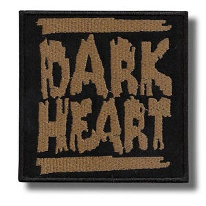 May include: A square embroidered patch featuring a black background with the words DARK HEART stitched in a textured, tan-coloured thread. The text is framed by two horizontal tan bars, one above and one below the lettering, with a black border.