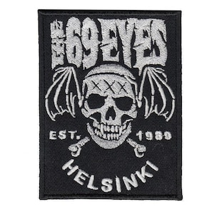 Può includere: Una toppa rettangolare nera ricamata con il testo bianco THE 69 EYES in alto. Sotto c'è un teschio bianco con ali di pipistrello e tre segni X sulla fronte. Il testo EST. 1989 e HELSINKI appare in ricamo bianco.
