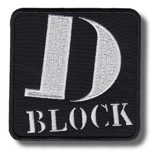 Può includere: Una toppa ricamata di forma quadrata con sfondo nero e bordo bianco. Il design presenta una grande lettera D bianca e la parola BLOCK ricamata in lettere maiuscole bianche sotto la lettera D sulla superficie in tessuto nero.