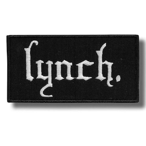 Puede incluir: Un parche rectangular negro bordado que presenta la palabra lynch. en letras blancas de estilo gótico. El texto está centrado sobre la tela, que tiene un borde cosido, creando un contraste claro entre el hilo blanco y el fondo negro del parche.