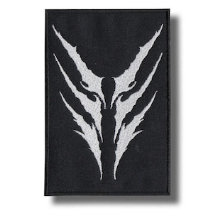 Puede incluir: Un parche rectangular bordado en negro con un diseño gráfico blanco simétrico. El diseño consiste en formas afiladas, dentadas y similares a garras que apuntan hacia arriba y hacia abajo, creando un emblema abstracto y estilizado.