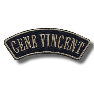 Puede incluir: Un parche bordado negro de forma curva que presenta el texto GENE VINCENT en letras mayúsculas de color tostado con serifa. El parche tiene un borde grueso cosido de color tostado que sigue la forma arqueada de la tela, creando un contorno.