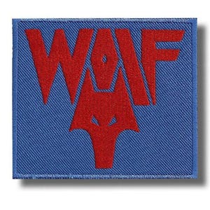 May include: A rectangular embroidered patch featuring a blue background with the red text WOLF. The letter O is stylised as a geometric wolf head icon, integrated into the centre of the design. The patch has a stitched border in matching blue.