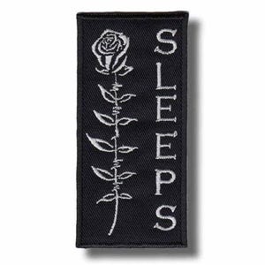 May include: A rectangular black embroidered patch featuring a white rose design on the left side and the word SLEEPS written vertically in white capital letters on the right side. The rose stem is depicted with a barbed wire texture.