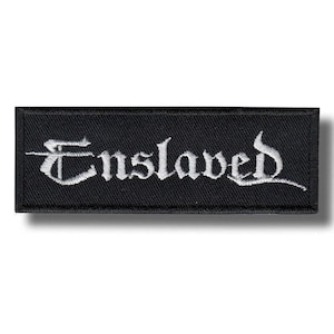 Puede incluir: Un parche rectangular bordado en negro con la palabra Enslaved cosida en hilo blanco. Las letras están representadas en una fuente de estilo gótico con adornos alargados en la primera y última letra, sobre un fondo de tela de color negro.