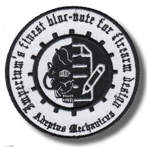 Puede incluir: Un parche bordado circular con un diseño en blanco y negro. El centro muestra un gráfico dividido de una calavera y un lápiz. El borde contiene el texto: Imperium's finest blue-note for firearm design Adeptus Mechanicus.