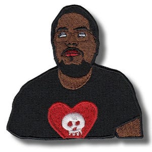 Pode incluir: Um emblema bordado apresentando uma pessoa de pele morena, com barba e cabelo escuro, vestindo uma camiseta preta. A camiseta exibe um gráfico de um grande coração vermelho contendo uma caveira branca com olhos vazios e nariz pequeno.