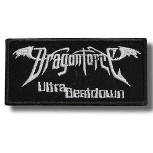 Puede incluir: Un parche rectangular negro bordado que presenta el texto blanco DragonForce en una fuente estilizada con detalles en forma de alas en los lados. Debajo del nombre de la banda, el texto Ultra Beatdown está bordado en una fuente blanca más pequeña.