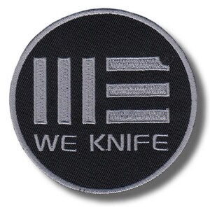 May include: A circular embroidered patch featuring a black background with a silver-grey border. The design includes three vertical silver-grey bars on the left, three horizontal silver-grey bars on the right, and the text WE KNIFE in silver-grey.