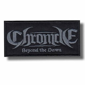 Op de afbeelding: Een rechthoekige zwarte geborduurde patch met het woord Chrome in een groot, gestileerd grijs gotisch lettertype, met de tekst Beyond the Dawn geborduurd in een kleiner, eenvoudiger grijs lettertype direct onder het hoofdwoord op de zwarte stof.