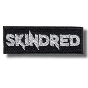 May include: A rectangular black embroidered patch featuring the word SKINDRED in bold, white, stylised capital letters. The text has a sharp, jagged font design with a thick black border surrounding the entire rectangular shape of the patch.