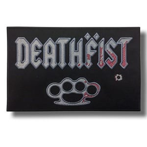 Op de afbeelding: Een rechthoekige zwarte geborduurde patch met het woord DEATHFIST in grijze blokletters met rode spetters. Onder de tekst staat een grijs geborduurd ontwerp van een boksbeugel met rode spetters en een klein kogelgat aan de rechterkant.