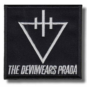 Op de afbeelding: Een vierkante zwarte geborduurde patch met een wit omgekeerd driehoekssymbool met een verticale lijn en twee kleinere horizontale balken erboven. De tekst THE DEVILWEARS PRADA is in witte hoofdletters langs de onderrand geborduurd.