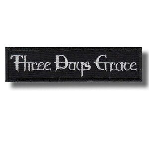 May include: A rectangular black embroidered patch featuring the text Three Days Grace in white, stylised, serif lettering. The patch has a clean, stitched border and a flat, fabric texture, presenting the band name in a clear, horizontal layout.