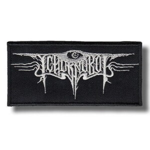 May include: A rectangular black embroidered patch featuring white thread detailing. The design displays stylised, jagged lettering that resembles a band logo, with an eye symbol centred above the text and sharp, pointed accents extending outwards.
