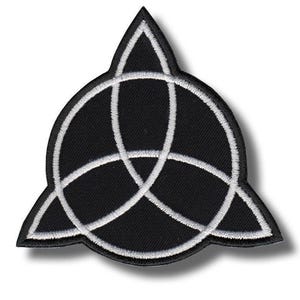 Op de afbeelding: Een zwarte geborduurde patch met een wit triquetra-symbool. Het ontwerp bestaat uit drie met elkaar verbonden bogen die een driehoekige vorm met puntige hoeken vormen, allemaal omlijnd met wit garen op een zwarte stoffen ondergrond.