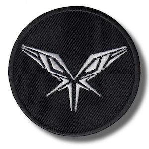 May include: A circular black embroidered patch featuring a white stylised wing or insect-like graphic centred on the fabric. The design consists of sharp, angular lines forming a symmetrical shape with a thick black stitched border edge.