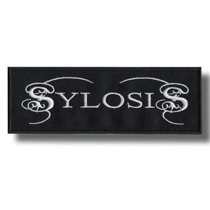 May include: A rectangular black embroidered patch featuring the word SYLOSIS in white capital letters. The letters S at the beginning and end of the word are stylized with decorative, swirling flourishes that extend outward from the main text.