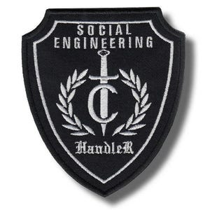 May include: A shield-shaped black embroidered patch with white stitching. The text reads SOCIAL ENGINEERING at the top and HandleR at the bottom. In the center, a white sword is positioned vertically behind a large letter C, flanked by laurel leaves.