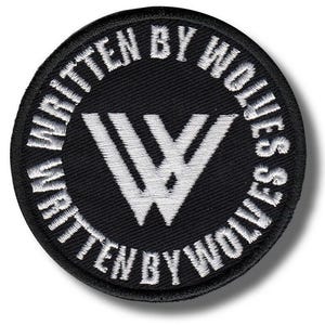 May include: A circular embroidered patch featuring a black background with white stitching. The center displays a stylized letter W, and the text WRITTEN BY WOLVES is stitched in a circular pattern around the border of the patch.