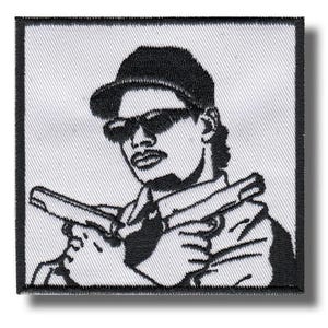 以下が含まれることがあります： 野球帽とサングラスを着用し、2丁の拳銃を構えた人物の白黒グラフィックイラストが刺繍された正方形のワッペン。白い布地を背景に、コントラストの強い黒い糸でデザインが表現されています。