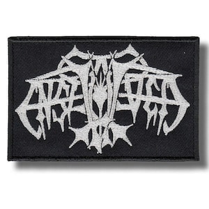 May include: A rectangular embroidered patch featuring a black background with a white, stylised, symmetrical logo design in the centre. The design consists of intricate, sharp, and jagged lines that form a complex, abstract emblem with a border.
