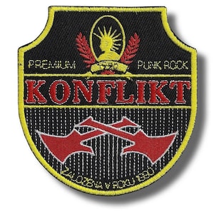 May include: A shield-shaped embroidered patch with a black background and yellow border. It features the red text KONFLIKT, the words PREMIUM PUNK ROCK, a yellow emblem of a figure with a spiked crown, and two crossed red arrows with white outlines.