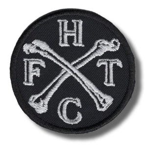 May include: A circular black embroidered patch featuring two crossed white bones in the centre. The letters H, F, T, and C are stitched in white thread, positioned between the bones in a clockwise arrangement starting from the top.