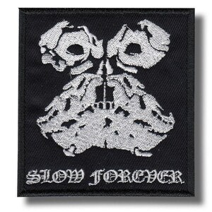 May include: A square black embroidered patch featuring a white graphic design of two symmetrical, abstract, skull-like shapes positioned above a textured, flowing form. The text SLOW FOREVER is embroidered in white in a gothic font at the bottom.