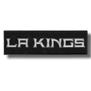 Pode incluir: Um emblema retangular bordado em preto com o texto LA KINGS em letras de bloco cinza-prateadas. O emblema possui uma borda costurada limpa e uma construção de tecido plano, projetada para aplicação em diversas superfícies têxteis.