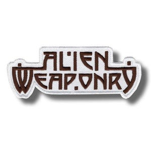 Puede incluir: Un parche bordado rectangular con fondo blanco y borde marrón oscuro. El texto ALIEN WEAPONRY está bordado con hilo marrón oscuro en una fuente angular estilizada. El parche tiene una forma distintiva con elementos laterales extendidos.