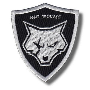May include: A shield-shaped embroidered patch featuring a black background with a silver-grey wolf head design. The wolf has a visible scar over one eye. The text "BAD WOLVES" is stitched in silver-grey thread above the wolf head design.