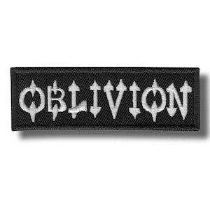 May include: A rectangular black embroidered patch featuring the word OBLIVION in a stylised white font. The letters have sharp, pointed edges and a distinct, angular design. The patch has a thick black border and a textured fabric surface.