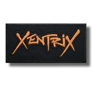 Puede incluir: Un parche rectangular bordado en negro que presenta la palabra XENTRIX cosida con una tipografía naranja estilizada y dentada. El parche tiene un borde acabado limpio y una superficie de tela texturizada, mostrando el nombre del grupo.