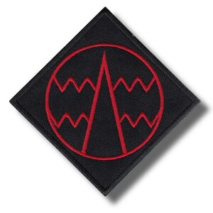 May include: A diamond-shaped black embroidered patch featuring a red circular border. Inside the circle, a red vertical triangle points upward, flanked by two horizontal red zigzag lines that extend from the triangle to the circular border.