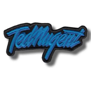 May include: An embroidered patch featuring the text TedMugent in a stylised, slanted blue script font. The lettering is outlined in a thick black border, creating a high-contrast design with a textured, stitched appearance throughout the surface.