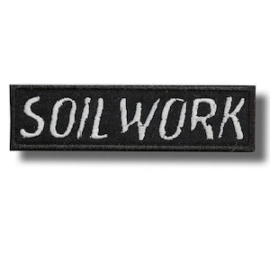 Puede incluir: Un parche rectangular bordado en negro con el texto SOIL WORK en letras mayúsculas blancas. El parche tiene un borde cosido limpio y una superficie de tela texturizada, diseñado para su aplicación en ropa u otros artículos textiles.