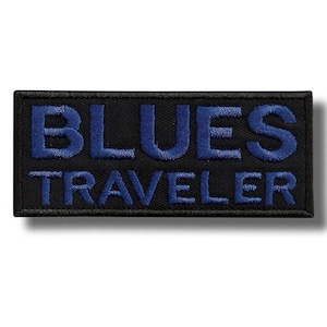 May include: A rectangular embroidered patch with a black background and a dark blue border. The text BLUES TRAVELER is stitched in a bold, dark blue sans-serif font across the center of the patch, arranged in two stacked lines.