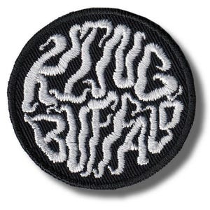 Puede incluir: Un parche bordado circular con fondo negro y hilo blanco que forma el texto "PLUG BUFFALO" en una fuente estilizada, ondulada e interconectada. Los bordes del parche circular están rematados con un denso borde cosido en color negro.