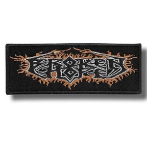 Puede incluir: Un parche rectangular bordado en negro que presenta la palabra BROKEN en una fuente estilizada y dentada de color blanco y plateado. El texto está rodeado por intrincados detalles de bordado en forma de llama naranja y marrón sobre fondo negro.