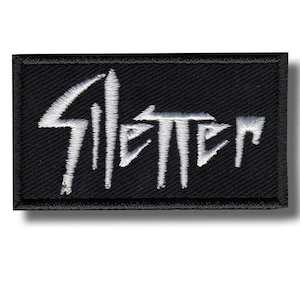 May include: A rectangular black embroidered patch featuring the word "Silencer" stitched in white thread with a stylized, sharp-edged font. The patch has a thick, stitched black border surrounding the entire rectangular design.