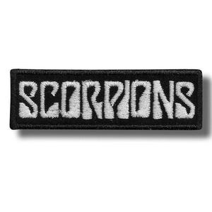 Puede incluir: Un parche rectangular bordado con fondo negro y la palabra SCORPIONS cosida en hilo blanco. Las letras presentan una fuente audaz y estilizada con un borde grueso, creando un diseño de alto contraste adecuado para aplicar sobre tela.