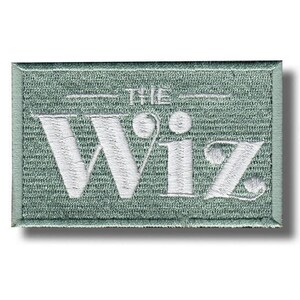 May include: A rectangular embroidered patch featuring a sage green background with the white text "THE WIZ" stitched in a stylised font. The patch has a matching sage green stitched border around the entire perimeter of the rectangular shape.