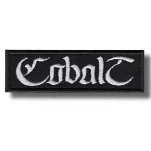 Op de afbeelding: Een rechthoekige geborduurde patch met een zwarte achtergrond en een dikke zwarte rand. Het woord Cobalt is in het midden geborduurd met witte draad in een gestileerd, gotisch lettertype met decoratieve krullen op de letters.